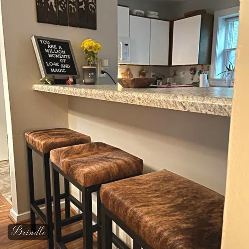 Peut inclure: Trois tabourets de bar en cuir de vachette brun et noir avec des cadres en m&eacute;tal noir. Les tabourets sont dans une cuisine avec un comptoir en granit et des armoires blanches. Une pancarte sur le comptoir indique "You are a million moments of love and magic."
