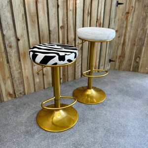 Op de afbeelding: Twee gouden barstoelen met verstelbare hoogte. E&eacute;n stoel heeft een wit kussen en de andere heeft een zwart-wit zebraprint kussen.