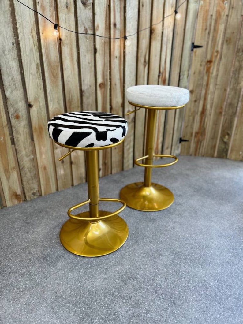 Op de afbeelding: Twee gouden barstoelen met verstelbare hoogte. E&eacute;n stoel heeft een wit kussen en de andere heeft een zwart-wit zebraprint kussen.