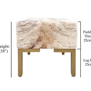 May include: A square ottoman with a cowhide top in shades of brown and cream, supported by gold-colored metal legs. The seat height is 18 inches, the padded top is 8.5 inches thick, and the leg height is 9 inches.