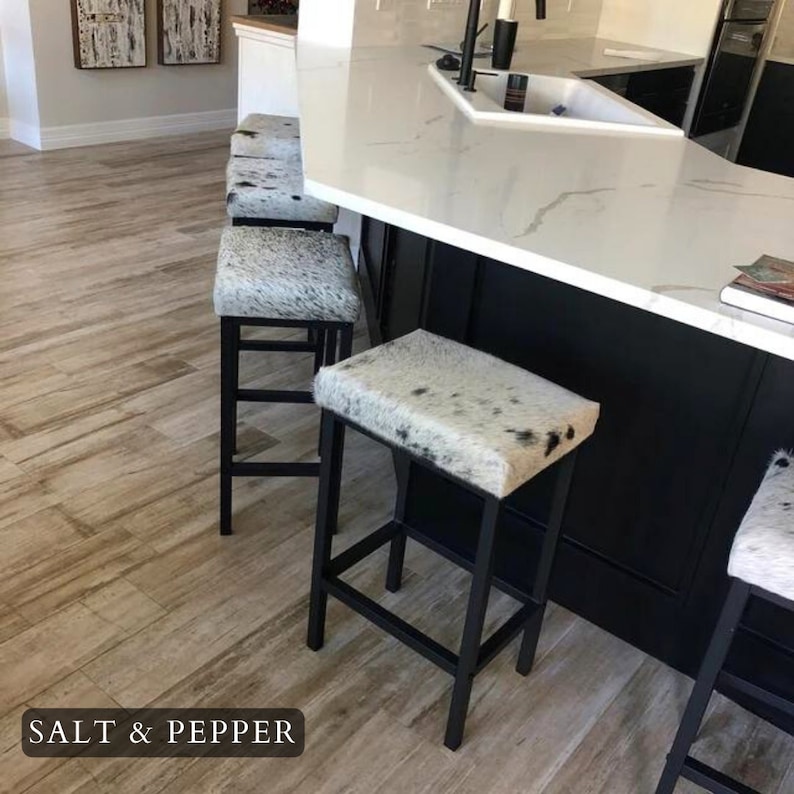 May include: Four black metal bar stools with white and black cowhide seats. The stools are arranged in a row in front of a kitchen island with a white countertop. The text "SALT & PEPPER" is visible on the floor.