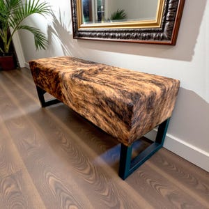 May include: A rectangular bench with a cowhide pattern in shades of brown and black. The bench has a black metal frame. The bench is placed in front of a wall with a mirror.