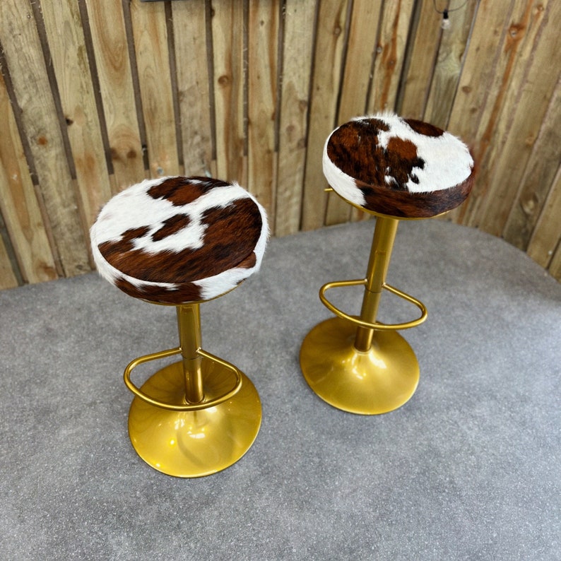 Op de afbeelding: Twee gouden barstoelen met bruine en witte koeienhuid zittingen. De krukken hebben een eenvoudig ontwerp met een ronde zitting en een enkele, verticale steun.