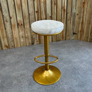 Op de afbeelding: Een gouden barstoel met een witte, gecapitonneerde zitting. De stoel heeft een ronde basis en een voetensteun.