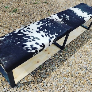 May include: A long black and white cowhide bench with a wooden base and black metal legs. The bench is sitting on a gravel surface.