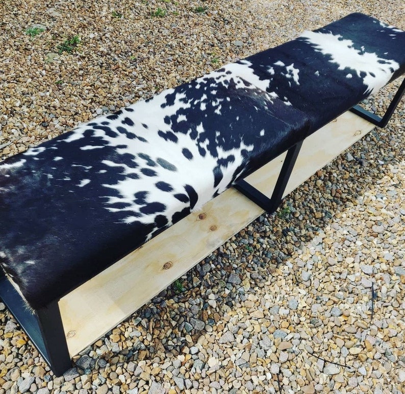 May include: A long black and white cowhide bench with a wooden base and black metal legs. The bench is sitting on a gravel surface.