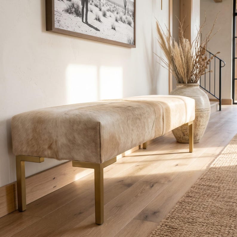 May include: A rectangular bench upholstered in light beige and white cowhide featuring a smooth, natural hair-on-hide texture. The bench is supported by four straight, metallic gold-toned legs with a clean, angular design.