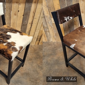 May include: Two black metal bar stools with a cowhide seat and back. The stool on the left has a tri-color cowhide pattern, while the stool on the right has a brown and white cowhide pattern. The stools are on a wooden floor.
