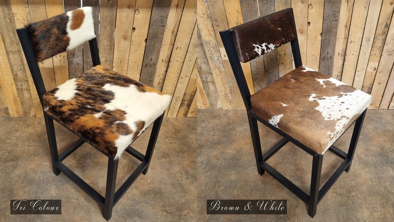 May include: Two black metal bar stools with a cowhide seat and back. The stool on the left has a tri-color cowhide pattern, while the stool on the right has a brown and white cowhide pattern. The stools are on a wooden floor.