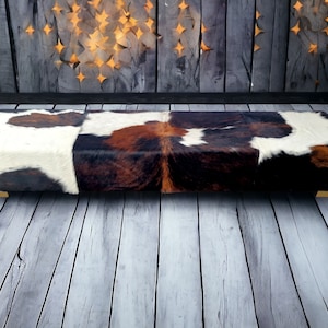 May include: A long, rectangular bench upholstered in a patchwork of brown, white, and black cowhide. The bench has a gold metal frame and sits on a light wood floor.
