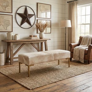 May include: A rectangular bench upholstered in light beige cowhide with a smooth texture, supported by four slender metallic gold legs. The bench is positioned on a woven jute rug in a room with white shiplap walls and wooden furniture.