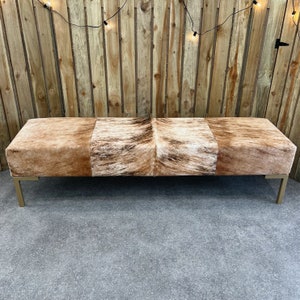 May include: A long, rectangular bench with a brown and white cowhide upholstery. The bench has a gold metal frame and is sitting on a gray concrete floor.
