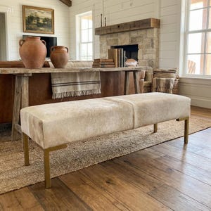 May include: A rectangular bench featuring a light beige, textured hide upholstery and four straight, metallic gold-toned legs. The bench is positioned horizontally in a room, showcasing a smooth, continuous top surface and a clean, structured frame design.