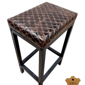 May include: A brown leather bar stool with a diamond stitch pattern. The stool has a black metal frame and a 100% genuine leather seat.