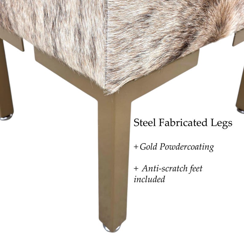 May include: Close-up of a square ottoman with a cowhide top in shades of brown and cream. The gold-colored steel legs have anti-scratch feet. The text on the image reads: "Steel Fabricated Legs + Gold Powdercoating + Anti-scratch feet included."
