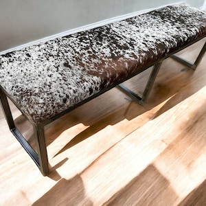 May include: A long, rectangular bench with a black metal frame and a brown and white cowhide seat.
