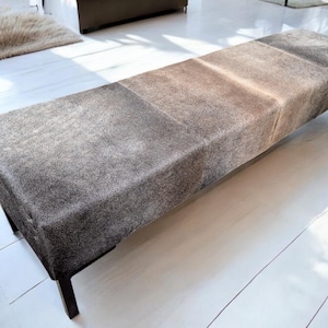 May include: A long, rectangular bench with a gray, brown, and white cowhide upholstery. The bench has a black metal frame and is sitting on a white wooden floor.