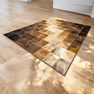 May include: A brown and beige patchwork cowhide rug with a dark brown border. The rug is made of squares of different shades of brown and beige cowhide.