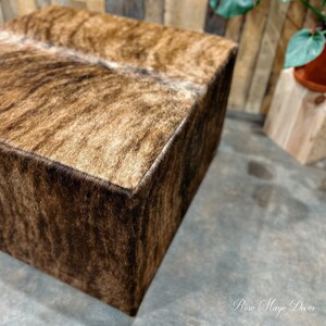 Pu&ograve; includere: Un pouf quadrato con un top in pelle di mucca marrone e bianca. Il pouf &egrave; in legno e ha una finitura naturale.