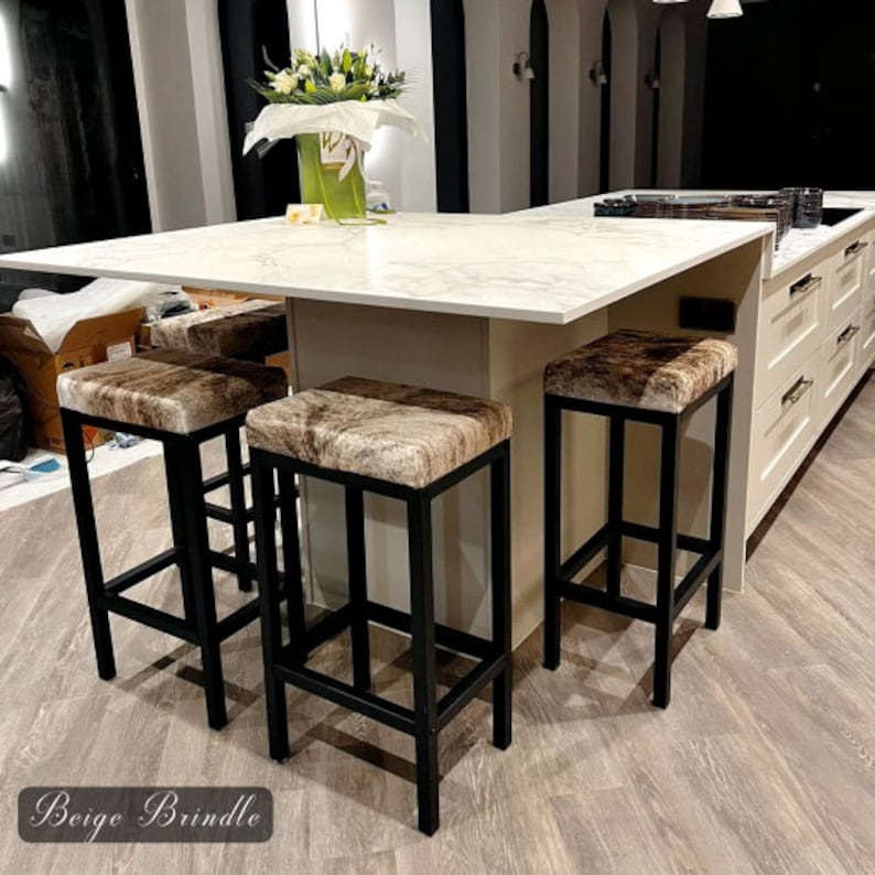 Peut inclure: Quatre tabourets de bar en m&eacute;tal noir avec des si&egrave;ges en peau de vache beige et marron. Les tabourets sont dispos&eacute;s autour d'un &icirc;lot de cuisine en marbre blanc.