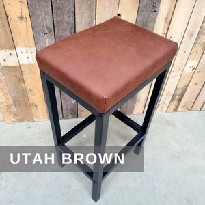 May include: A brown leather bar stool with a black metal frame. The stool has a square seat and a simple design. The text "UTAH BROWN" is visible in the image.
