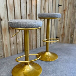 Op de afbeelding: Twee gouden barstoelen met grijze stoffen zittingen. De krukken hebben een ronde basis en een voetensteun.