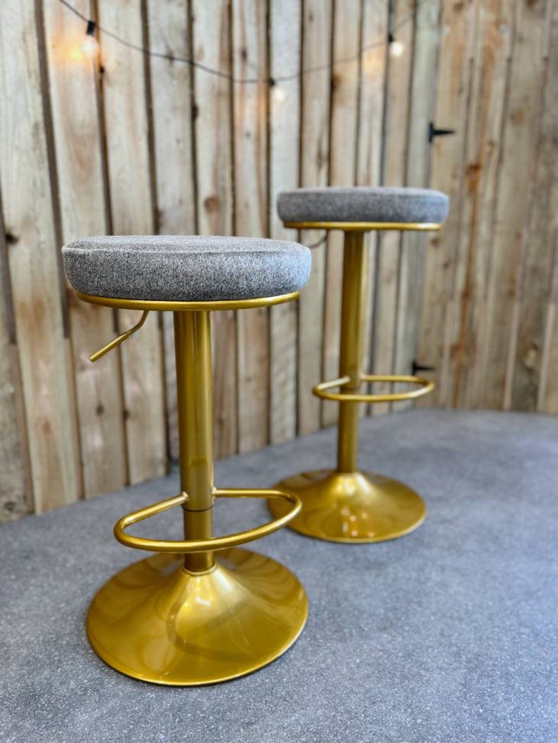 Op de afbeelding: Twee gouden barstoelen met grijze stoffen zittingen. De krukken hebben een ronde basis en een voetensteun.