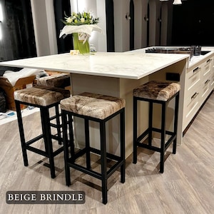 May include: Four black metal bar stools with beige and brown cowhide seats. The stools are arranged around a white marble kitchen island.