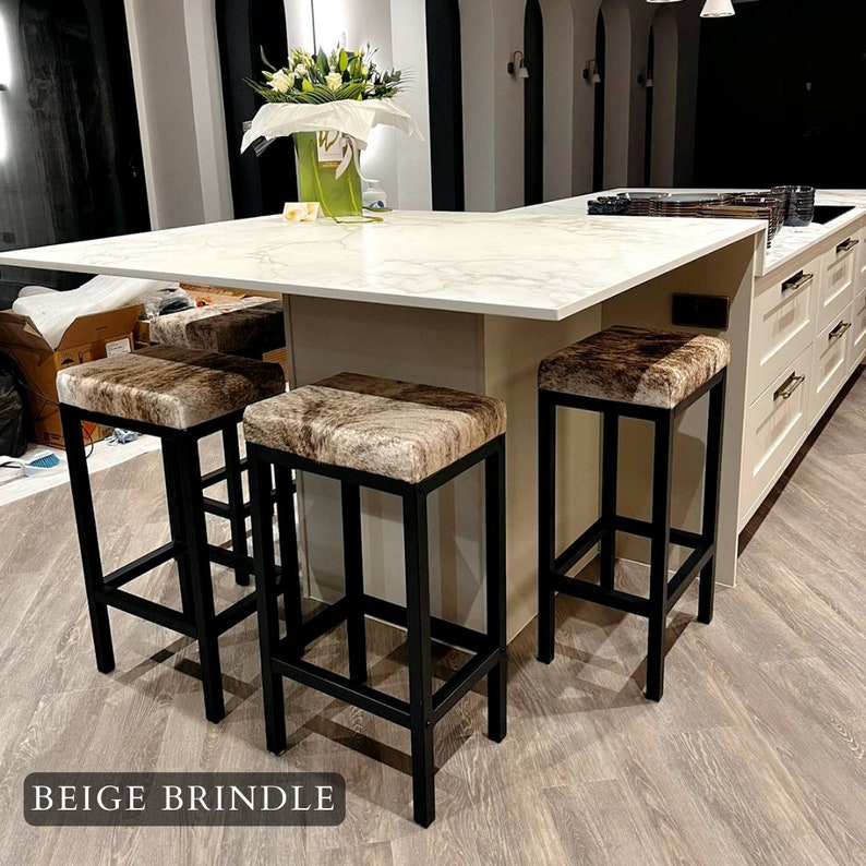 May include: Four black metal bar stools with beige and brown cowhide seats. The stools are arranged around a white marble kitchen island.