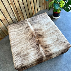 May include: A square, brown and white cowhide ottoman with a natural, textured surface.