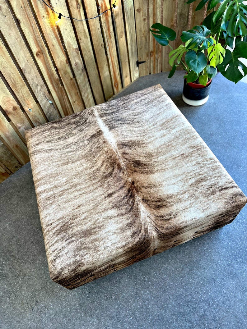 May include: A square, brown and white cowhide ottoman with a natural, textured surface.