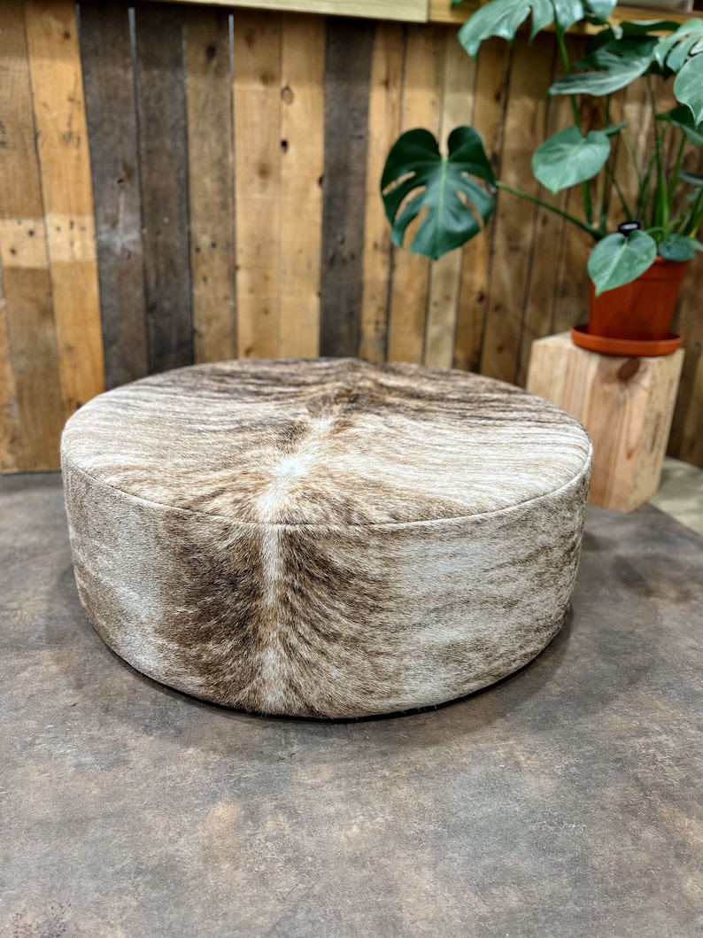 May include: A round, brown and white cowhide ottoman with a soft, plush texture. The ottoman is sitting on a brown floor.
