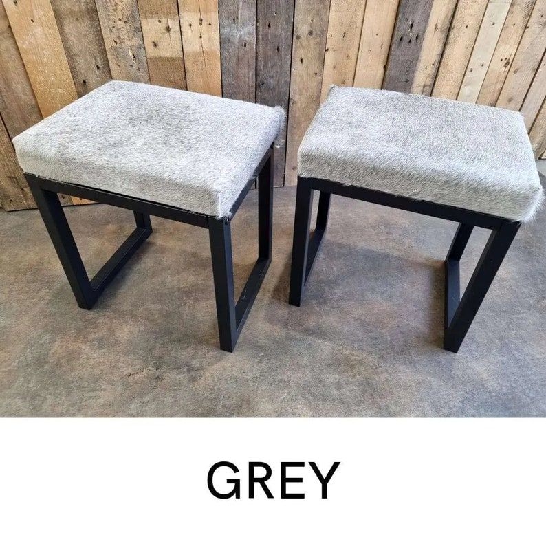 Puede incluir: Dos taburetes de piel de vaca gris con marcos de metal negro. Los taburetes est&aacute;n sobre un suelo de hormig&oacute;n.