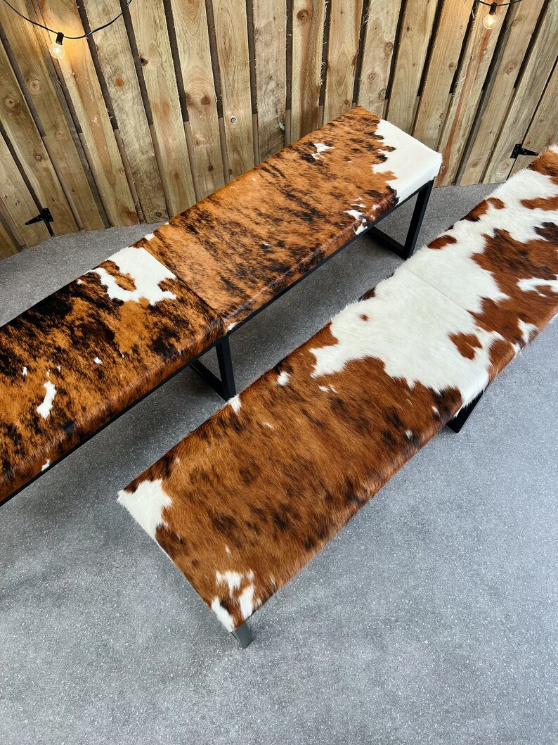 May include: Two brown and white cowhide benches with black metal legs. The benches are on a gray concrete floor.