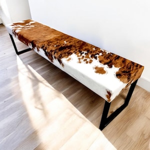 May include: A long, rectangular bench with a white and brown cowhide upholstered seat and black metal legs.