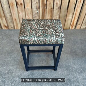 Op de afbeelding: Een zwarte metalen kruk met een vierkante zitting bekleed met een bloemenstof in turquoise en bruine tinten. De stof heeft een reli&euml;fpatroon.