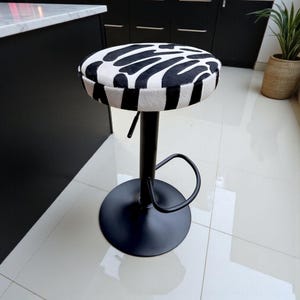 May include: A black and white zebra print bar stool. The round seat features a bold black and white pattern. The stool has a black, adjustable-height stem and a circular base. A curved footrest is attached to the stem.