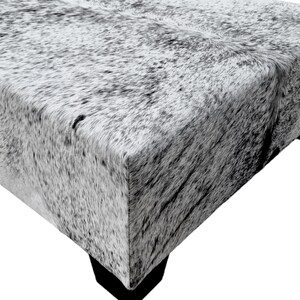 May include: A gray and white cowhide ottoman with a square top and four legs. The ottoman is made of real cowhide and has a natural, rustic look.