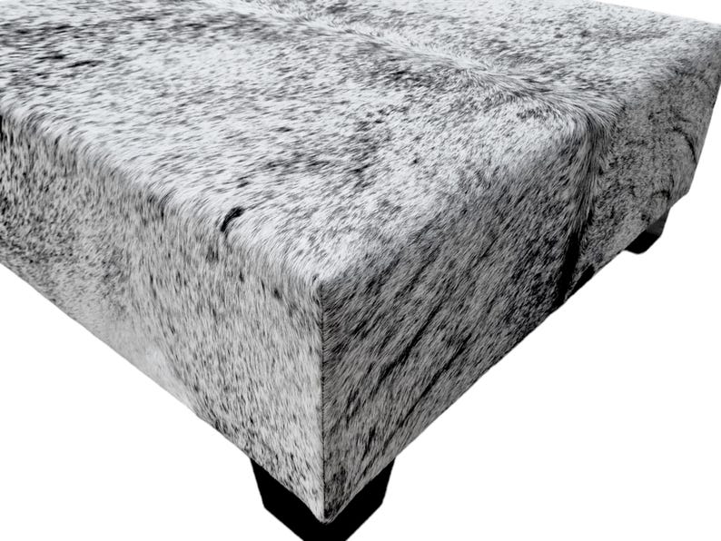 May include: A gray and white cowhide ottoman with a square top and four legs. The ottoman is made of real cowhide and has a natural, rustic look.