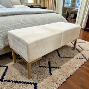 May include: A rectangular bench with a cream-colored, textured surface. The bench has a light brown metal frame and four legs. The bench is placed on a beige rug with a dark blue geometric pattern. The bench is in front of a bed.