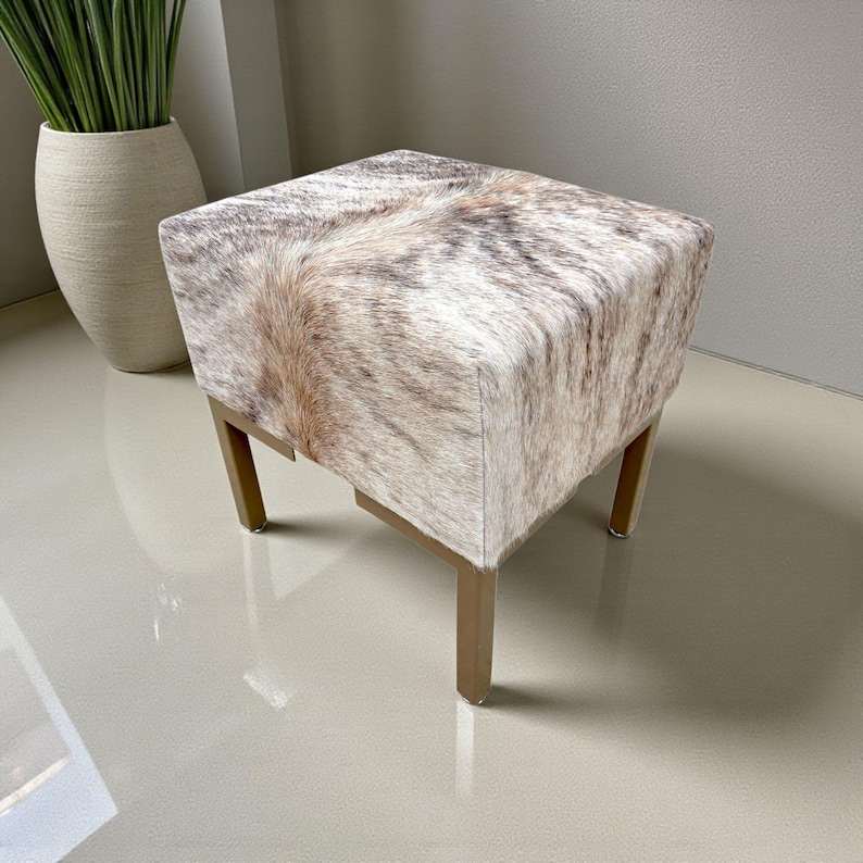 May include: A square ottoman with a cowhide pattern in shades of brown, tan, and white. The ottoman has a wooden frame with four legs. The design is a mix of natural and neutral tones, suitable for various interior styles.