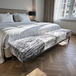 May include: A modern bench with a black metal frame and three cushioned seats. The seats are upholstered in a black, white, and gray patterned fabric, resembling animal hide. The bench is placed at the foot of a bed with white linens.