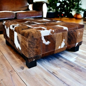 May include: A brown and white patchwork cowhide ottoman with a square top and four black legs. The ottoman is sitting on a wooden floor.
