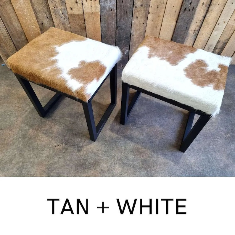 Puede incluir: Dos peque&ntilde;os taburetes cuadrados de metal negro con tapicer&iacute;a de piel de vaca marr&oacute;n y blanca. TAN + WHITE est&aacute; escrito en blanco sobre fondo negro.