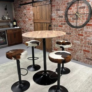Op de afbeelding: Een ronde houten tafel met een zwarte metalen basis en vier barkrukken van koeienhuid. De krukken hebben zwarte metalen bases en voetsteunen. De tafel en krukken staan in een kamer met een bakstenen muur en een houten deur.