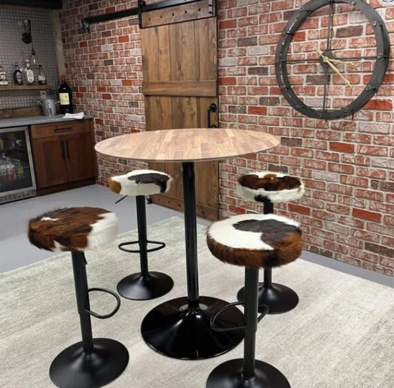 Op de afbeelding: Een ronde houten tafel met een zwarte metalen basis en vier barkrukken van koeienhuid. De krukken hebben zwarte metalen bases en voetsteunen. De tafel en krukken staan in een kamer met een bakstenen muur en een houten deur.