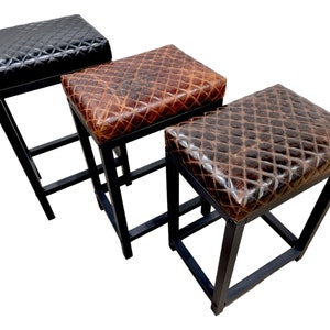 May include: Three black metal bar stools with different colored leather seats. The seats are all quilted and have a distressed look. The stools are all the same height and have a simple, modern design.