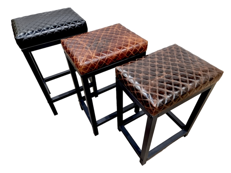 May include: Three black metal bar stools with different colored leather seats. The seats are all quilted and have a distressed look. The stools are all the same height and have a simple, modern design.
