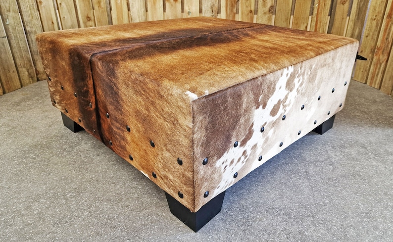 May include: A square brown and white cowhide ottoman with black metal studs and black wooden legs.