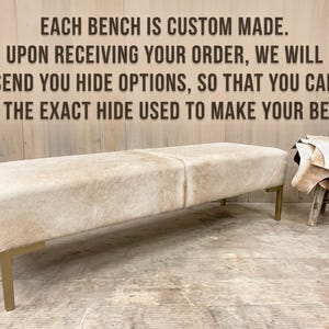 May include: A rectangular bench upholstered in light beige cowhide with a smooth texture, supported by four metallic gold-toned legs. The text reads: EACH BENCH IS CUSTOM MADE. UPON RECEIVING YOUR ORDER, WE WILL SEND YOU HIDE OPTIONS.